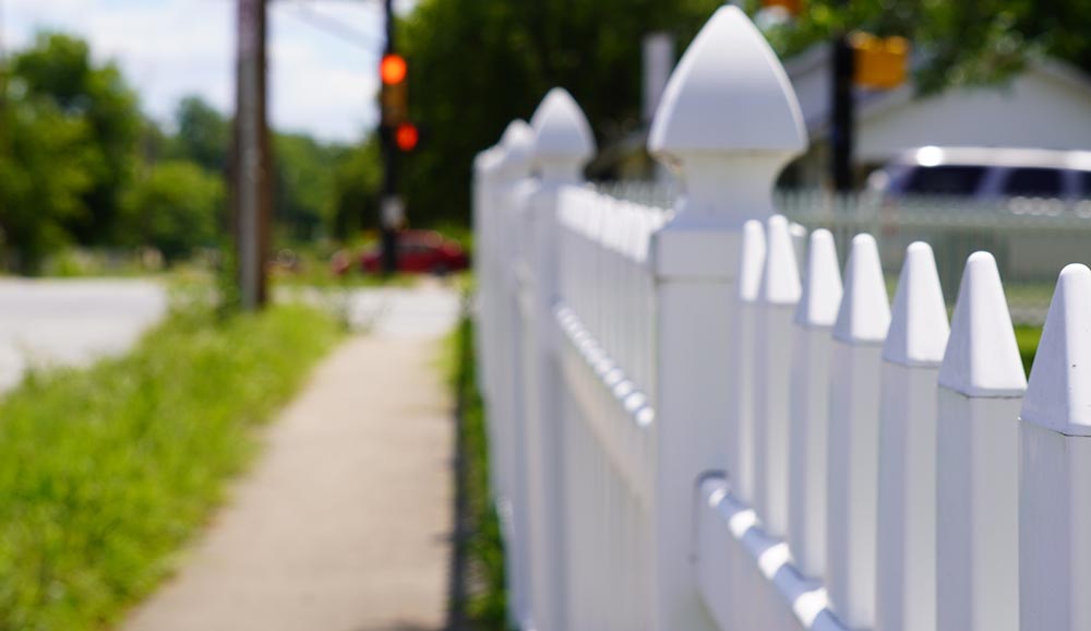 Fence Line Landscaping, a white picket fence along a sidewalk and grass between the sidewalk and the street.